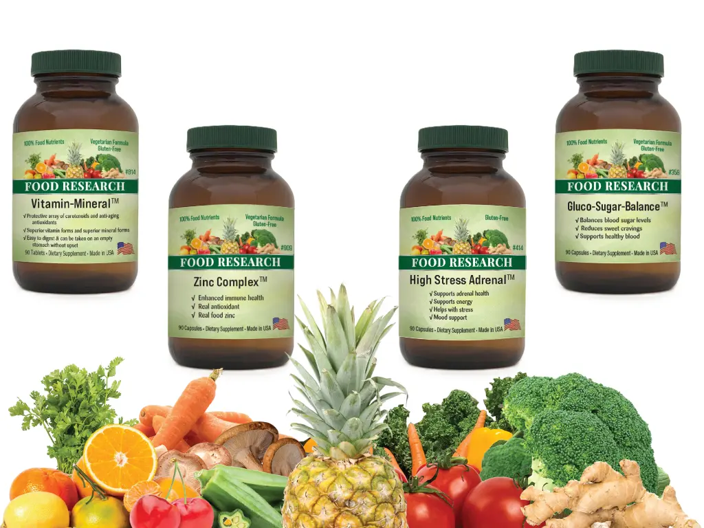 Four amber Food Research supplement bottles (Vitamin-Mineral, Zinc Complex, High Stress Adrenal, Gluco-Sugar-Balance) with green caps, displayed above a colorful assortment of fresh fruits and vegetables including pineapple, oranges, broccoli, carrots, and ginger.